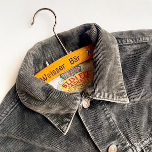 Op de afbeelding: Close-up van een donkergrijze corduroy jas met een knoopsluiting. De jas hangt aan een houten kleerhanger met de tekst "Weisser Bär" en een Levi's label. De jas heeft twee voorzakken en een kraag.