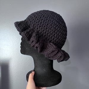 May include: A black crochet hat with a ruffled brim and a decorative flower on the side. The hat is displayed on a black mannequin head against a gray background. The hat is handmade and has a textured appearance.