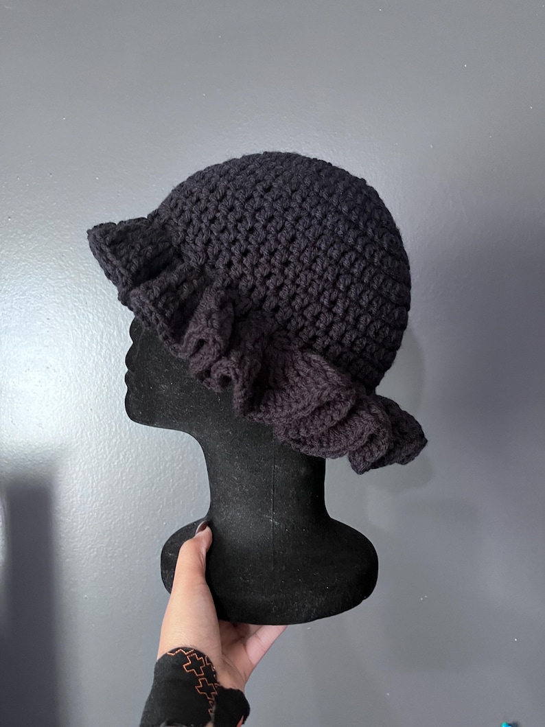 May include: A black crochet hat with a ruffled brim and a decorative flower on the side. The hat is displayed on a black mannequin head against a gray background. The hat is handmade and has a textured appearance.