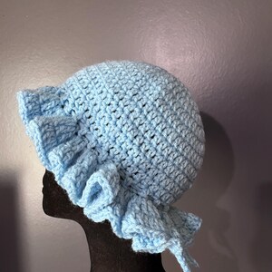 May include: A light blue, handmade crochet bonnet with a ruffled brim. The bonnet is made of soft yarn and features a rounded crown and ties for securing. The bonnet is displayed on a black mannequin head against a gray background.