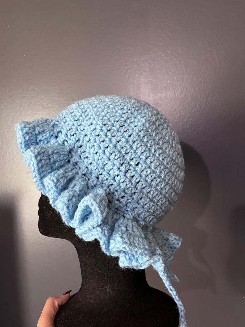 May include: A light blue, handmade crochet bonnet with a ruffled brim. The bonnet is made of soft yarn and features a rounded crown and ties for securing. The bonnet is displayed on a black mannequin head against a gray background.
