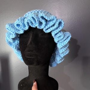 May include: A light blue, crocheted bonnet with a ruffled edge. The bonnet is displayed on a black mannequin head against a gray background. The handmade hat is a soft, pastel blue color.