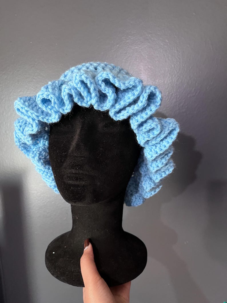May include: A light blue, crocheted bonnet with a ruffled edge. The bonnet is displayed on a black mannequin head against a gray background. The handmade hat is a soft, pastel blue color.
