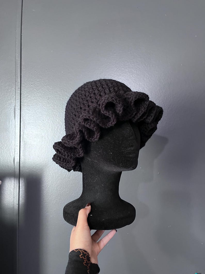 May include: A black crochet hat with a ruffled brim is displayed on a black mannequin head. The hat is made of a thick, textured yarn, and the ruffled brim adds a decorative element. The hat is a dark shade of black.
