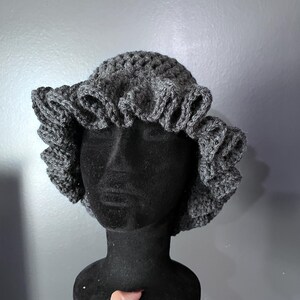 May include: A gray crochet hat with a ruffled brim is displayed on a black mannequin head. The hat is made of a textured yarn and has a rounded crown. The ruffled brim adds a decorative element to the hat.