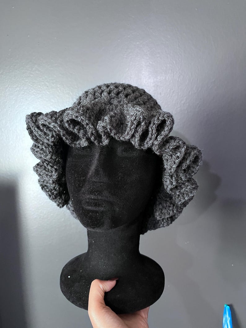 May include: A gray crochet hat with a ruffled brim is displayed on a black mannequin head. The hat is made of a textured yarn and has a rounded crown. The ruffled brim adds a decorative element to the hat.