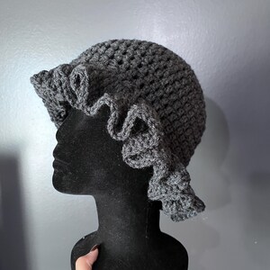 May include: A dark gray, crocheted hat with a ruffled brim. The hat is displayed on a black mannequin head. The crochet work has a textured appearance, and the ruffled edge adds a decorative element to the hat's design.