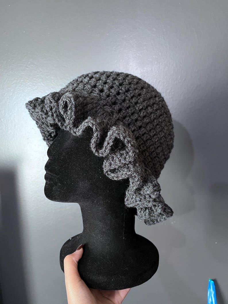 May include: A dark gray, crocheted hat with a ruffled brim. The hat is displayed on a black mannequin head. The crochet work has a textured appearance, and the ruffled edge adds a decorative element to the hat's design.