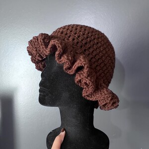 May include: A brown crochet hat with a ruffled brim. The hat is displayed on a black mannequin head against a gray background. The hat's texture is visible, showcasing the handmade quality.