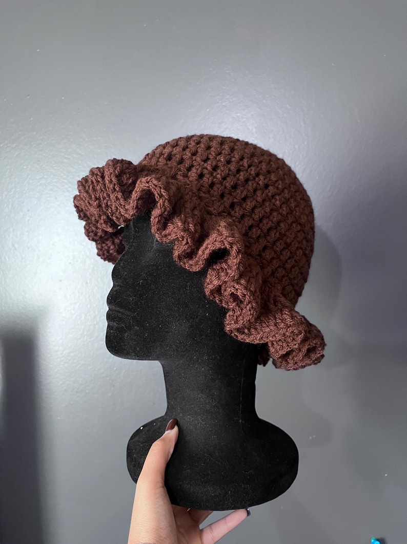 May include: A brown crochet hat with a ruffled brim. The hat is displayed on a black mannequin head against a gray background. The hat's texture is visible, showcasing the handmade quality.