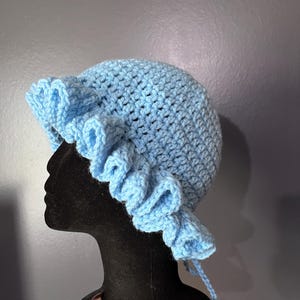 May include: A light blue crochet hat with a ruffled brim. The hat is displayed on a black mannequin head. The hat is handmade and has a textured appearance. The hat has a rounded crown and a wide brim with a decorative ruffle. The hat is tied with a string.