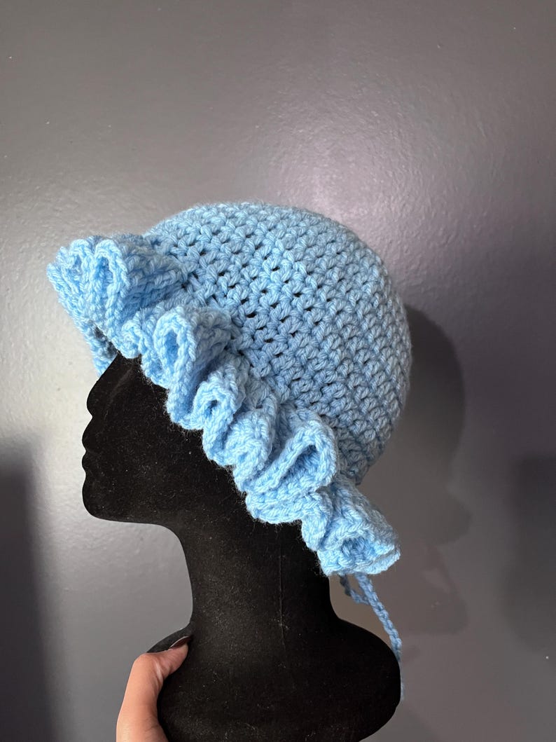 May include: A light blue crochet hat with a ruffled brim. The hat is displayed on a black mannequin head. The hat is handmade and has a textured appearance. The hat has a rounded crown and a wide brim with a decorative ruffle. The hat is tied with a string.