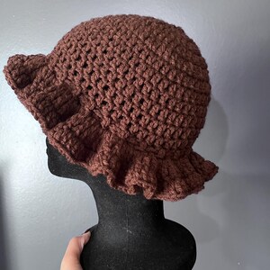 May include: A brown crochet hat with a ruffled brim. The hat is displayed on a black mannequin head against a gray background. The hat's texture is visible, showcasing the handmade crochet stitches.
