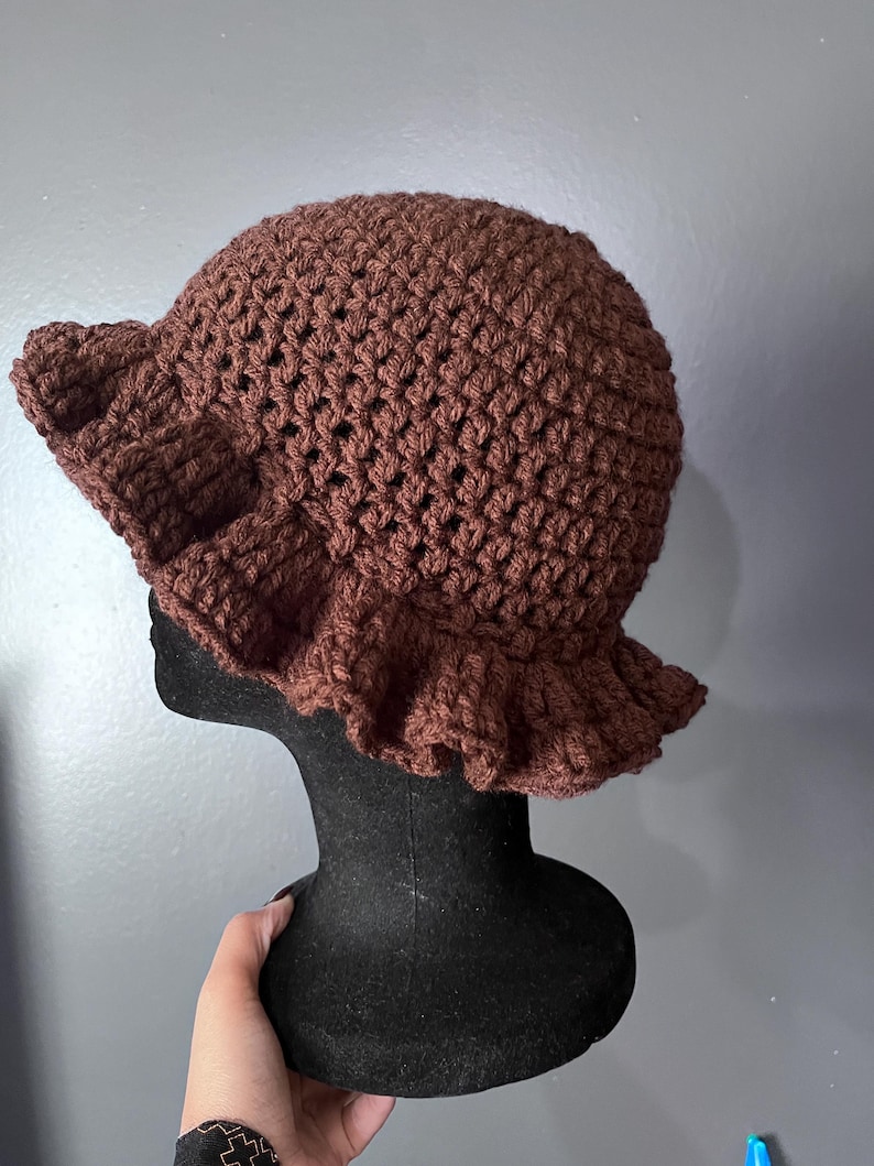 May include: A brown crochet hat with a ruffled brim. The hat is displayed on a black mannequin head against a gray background. The hat's texture is visible, showcasing the handmade crochet stitches.