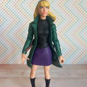 May include: A green jacket with a black turtleneck and purple skirt. The figure has blonde hair and is wearing black boots.
