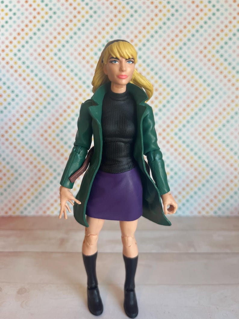 May include: A green jacket with a black turtleneck and purple skirt. The figure has blonde hair and is wearing black boots.
