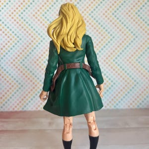 May include: A green and brown action figure of a woman with long blonde hair. She is wearing a green coat, a brown belt, and black shoes. The figure is posed with her arms at her sides and her legs slightly apart.