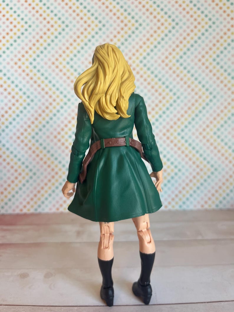 May include: A green and brown action figure of a woman with long blonde hair. She is wearing a green coat, a brown belt, and black shoes. The figure is posed with her arms at her sides and her legs slightly apart.