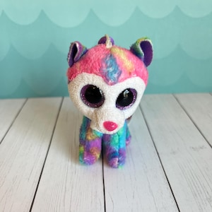 May include: A colorful plush toy animal with large, glittery eyes and a pink nose. The toy features a white face and a body with rainbow-colored fur. The ears are purple and green. The toy is standing on a white wooden surface.