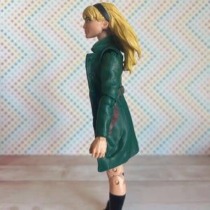 May include: A green and brown action figure of a woman with blonde hair wearing a green coat and black boots.