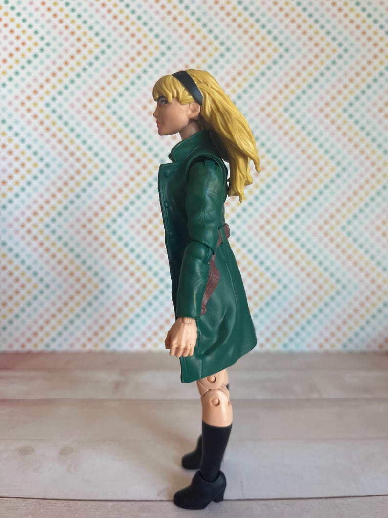 May include: A green and brown action figure of a woman with blonde hair wearing a green coat and black boots.