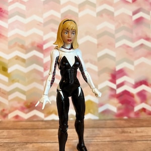 Peut inclure: Une figurine d'action blanche et noire d'une super-h&eacute;ro&iuml;ne f&eacute;minine aux cheveux blonds. Elle porte une combinaison blanche et noire avec un symbole d'araign&eacute;e sur la poitrine. La figurine est debout sur une surface en bois.