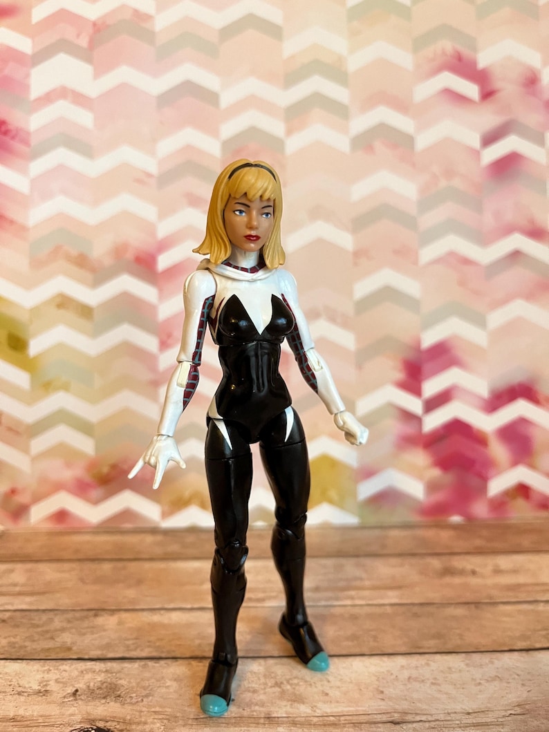 Peut inclure: Une figurine d'action blanche et noire d'une super-h&eacute;ro&iuml;ne f&eacute;minine aux cheveux blonds. Elle porte une combinaison blanche et noire avec un symbole d'araign&eacute;e sur la poitrine. La figurine est debout sur une surface en bois.