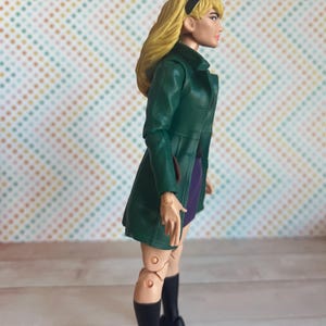 May include: A green and purple action figure of a woman with blonde hair wearing a green coat and black boots.