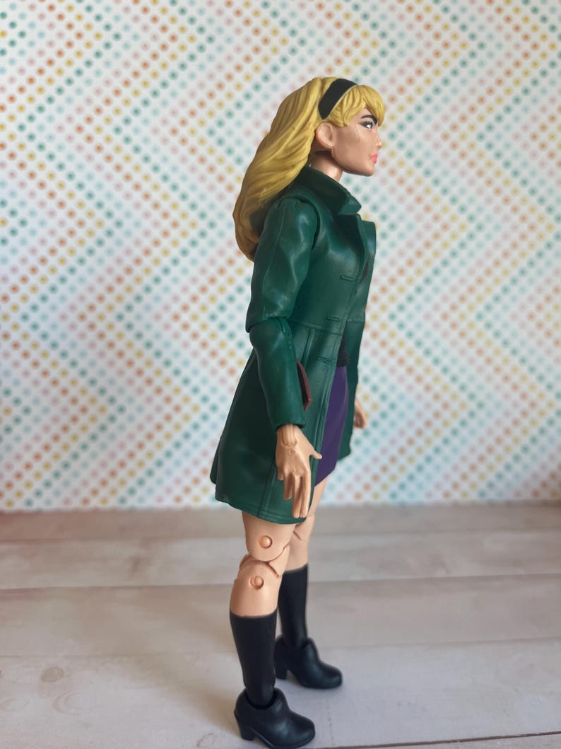 May include: A green and purple action figure of a woman with blonde hair wearing a green coat and black boots.