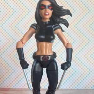 May include: A black and grey action figure of a female superhero with red eye coverings. The figure is holding two silver swords in each hand.