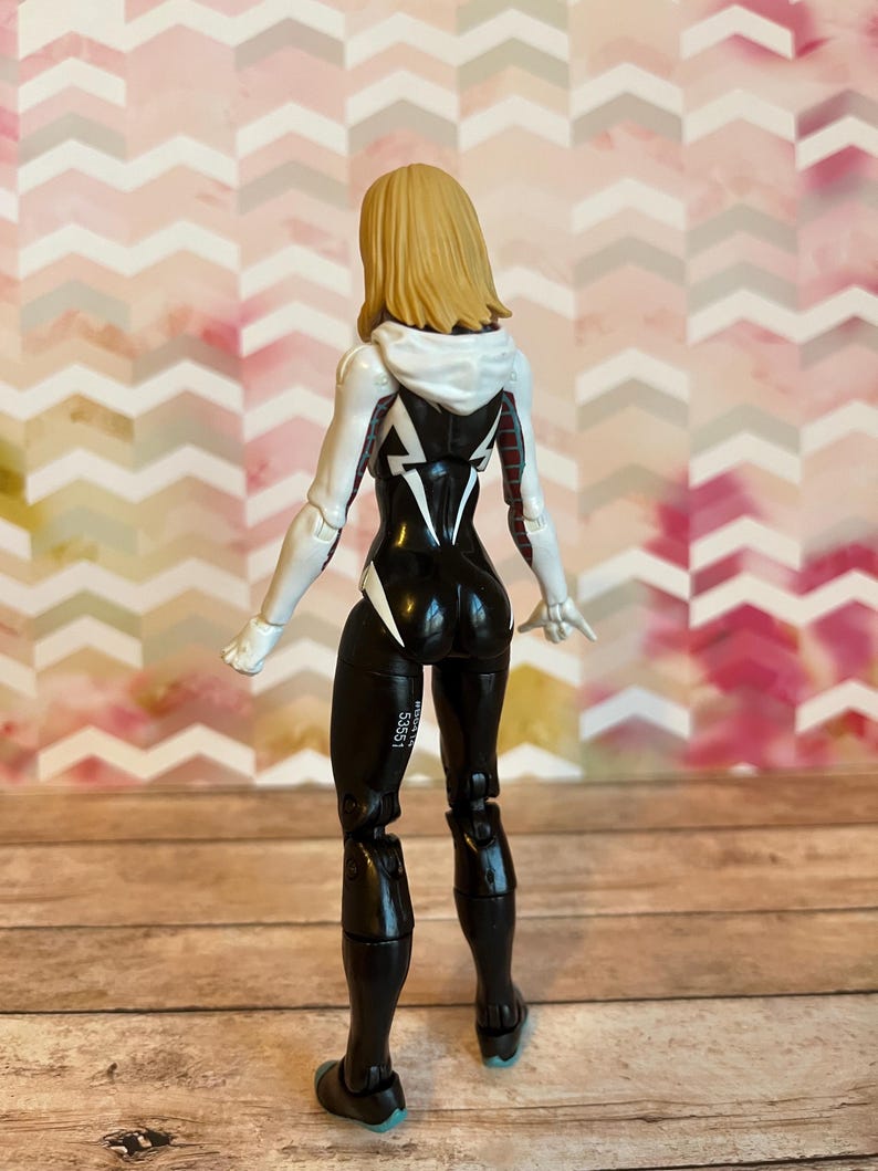Peut inclure: Une figurine d'action noire et blanche d'une super-h&eacute;ro&iuml;ne f&eacute;minine aux longs cheveux blonds. La figurine porte une veste &agrave; capuche blanche avec des accents noirs et rouges. La figurine est debout, dos &agrave; la cam&eacute;ra.