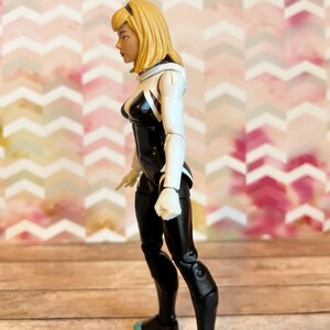 Peut inclure: Une figurine d'action noire et blanche d'une super-h&eacute;ro&iuml;ne f&eacute;minine aux cheveux blonds. Elle porte une combinaison blanche et noire avec un symbole d'araign&eacute;e sur la poitrine. La figurine est pos&eacute;e avec une jambe en avant et les bras le long du corps.