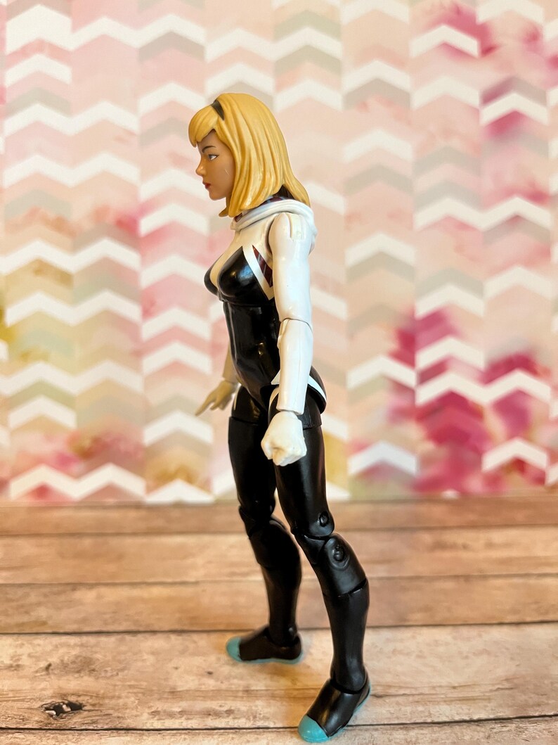 Peut inclure: Une figurine d'action noire et blanche d'une super-h&eacute;ro&iuml;ne f&eacute;minine aux cheveux blonds. Elle porte une combinaison blanche et noire avec un symbole d'araign&eacute;e sur la poitrine. La figurine est pos&eacute;e avec une jambe en avant et les bras le long du corps.