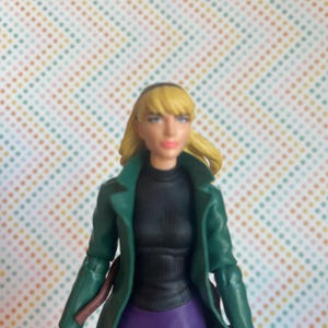 May include: A green jacket with a black turtleneck and purple skirt. The figure has blonde hair and is wearing a black headband.