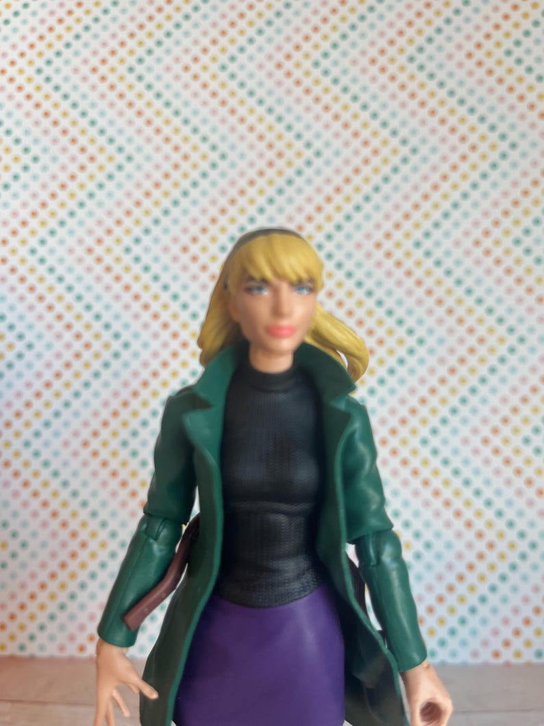 May include: A green jacket with a black turtleneck and purple skirt. The figure has blonde hair and is wearing a black headband.