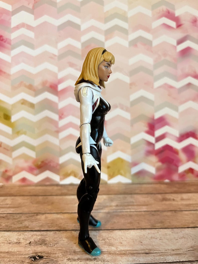 Peut inclure: Une figurine d'action blanche et noire d'une super-h&eacute;ro&iuml;ne avec des cheveux blonds. La figurine est pos&eacute;e avec un bras lev&eacute; et l'autre bras pli&eacute; au coude. La figurine porte une combinaison blanche et noire avec un symbole d'araign&eacute;e sur la poitrine.