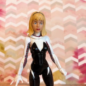 Peut inclure: Une figurine d'action blanche et noire d'une super-h&eacute;ro&iuml;ne f&eacute;minine aux cheveux blonds. La figurine porte une combinaison noire et blanche avec un symbole d'araign&eacute;e blanc sur la poitrine. La figurine est pos&eacute;e avec un bras lev&eacute; et l'autre bras tendu.