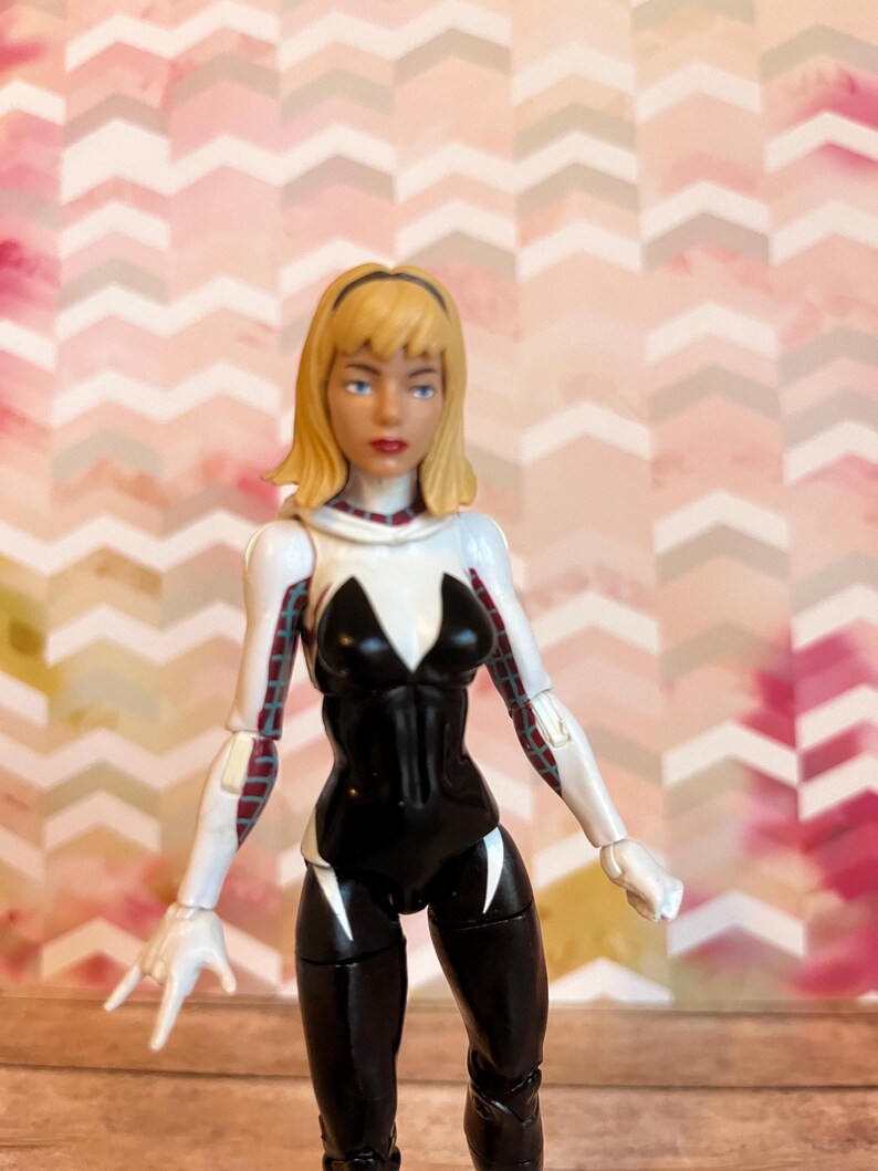 Peut inclure: Une figurine d'action blanche et noire d'une super-h&eacute;ro&iuml;ne f&eacute;minine aux cheveux blonds. La figurine porte une combinaison noire et blanche avec un symbole d'araign&eacute;e blanc sur la poitrine. La figurine est pos&eacute;e avec un bras lev&eacute; et l'autre bras tendu.