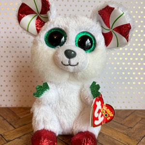 May include: A white plush toy mouse with large green eyes and candy cane ears. The mouse has red sequined feet and a red heart-shaped tag. The toy is a festive holiday item.