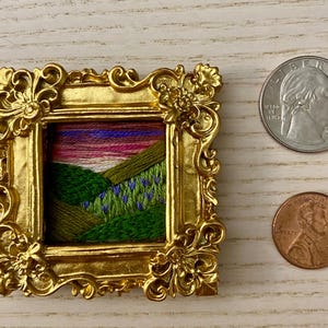 May include: A small embroidered landscape scene of a green hillside with purple flowers, framed in a gold ornate frame.