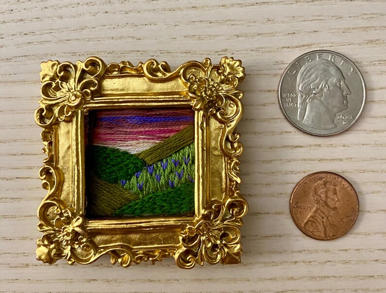 May include: A small embroidered landscape scene of a green hillside with purple flowers, framed in a gold ornate frame.