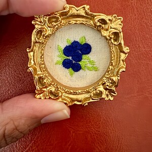 May include: A gold-colored, ornate frame with a small, embroidered picture of blueberries. The blueberries are a deep blue color, and the leaves are green. The frame is held by a hand against a red background.