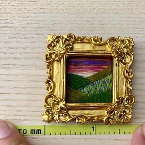 May include: A small, gold-colored picture frame with a colorful embroidered landscape scene inside. The frame is ornate and has a detailed design. The scene is a green field with purple flowers and a pink and orange sunset in the background.