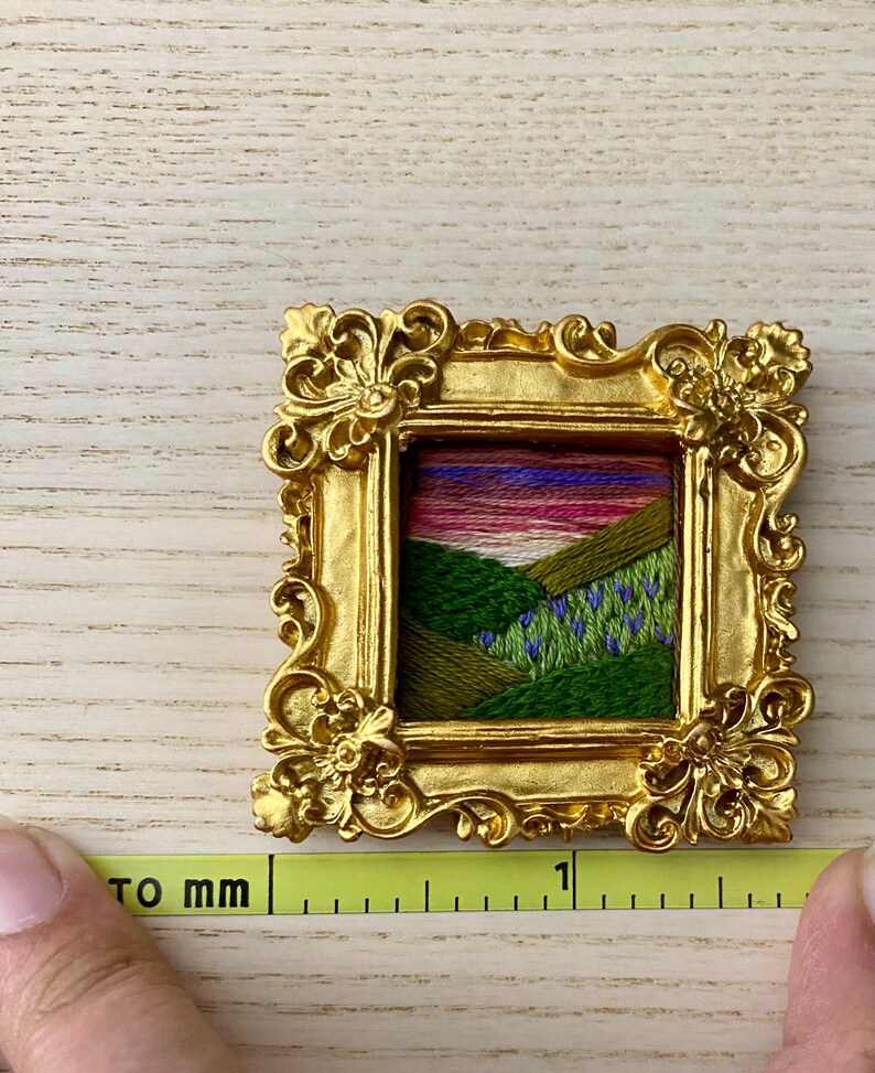 May include: A small, gold-colored picture frame with a colorful embroidered landscape scene inside. The frame is ornate and has a detailed design. The scene is a green field with purple flowers and a pink and orange sunset in the background.
