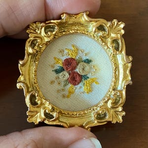 May include: A small, round, embroidered picture of flowers in a gold frame. The flowers are red, white, and yellow.