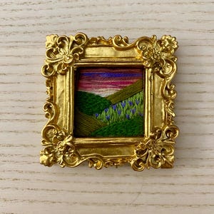 May include: A small embroidered landscape scene of a green hillside with purple flowers under a pink and purple sunset, framed in a gold ornate frame.