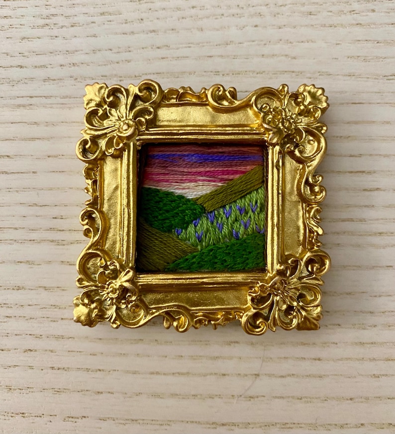 May include: A small embroidered landscape scene of a green hillside with purple flowers under a pink and purple sunset, framed in a gold ornate frame.
