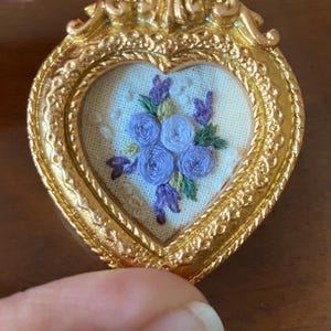 May include: A small heart-shaped gold frame with a delicate floral embroidery of purple and white flowers. The embroidery is framed in a gold heart-shaped frame with ornate details.