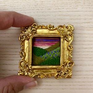 May include: A small framed embroidery depicting a landscape with green hills, a winding path, and a purple sunset. The frame is gold and ornate.