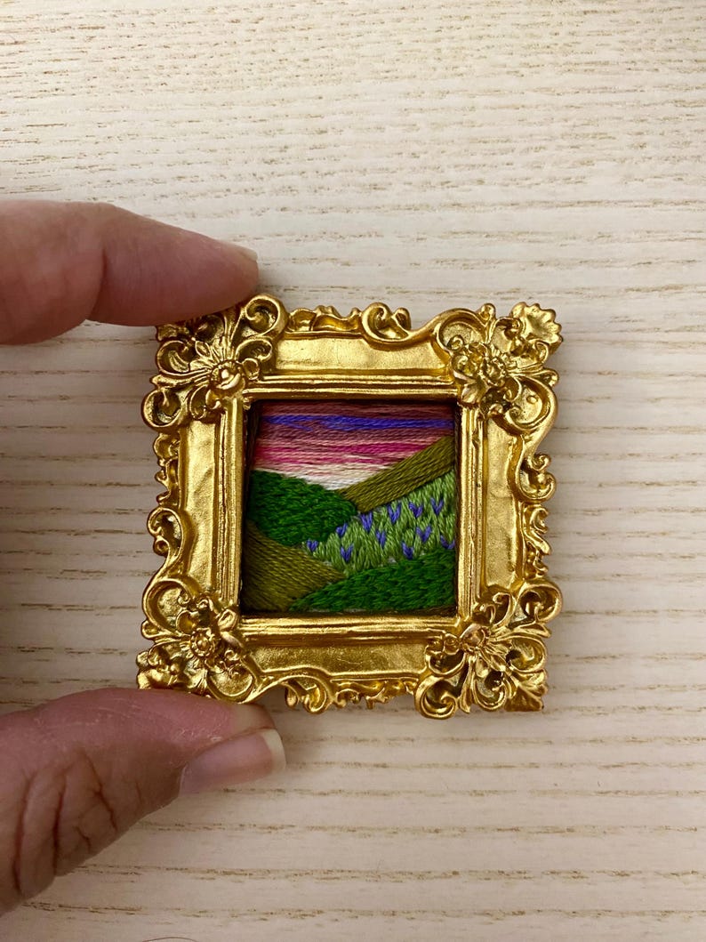 May include: A small framed embroidery depicting a landscape with green hills, a winding path, and a purple sunset. The frame is gold and ornate.